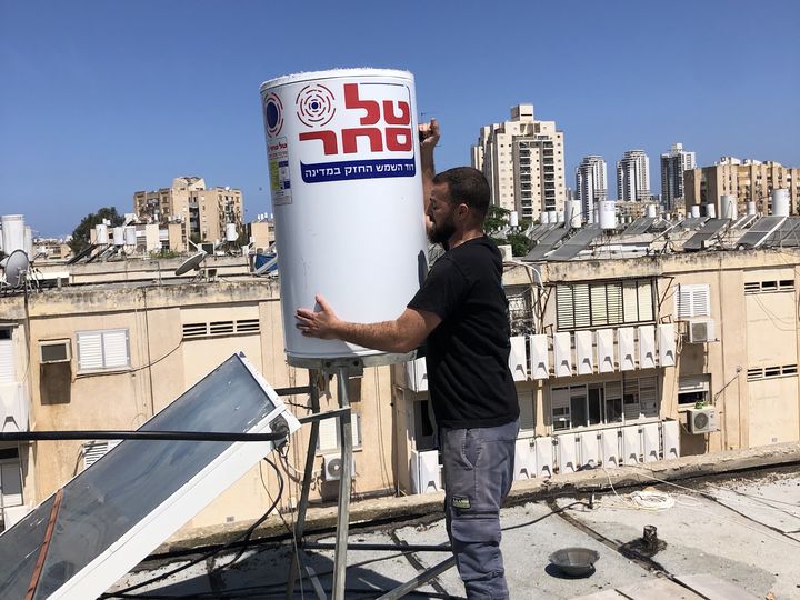 איש מקצוע מציב דוד מים לבן על מתקן על גג בניין, ברקע בניינים ושמיים כחולים